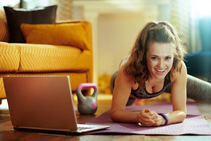 smiling fit woman in sport clothes in the modern house using online streaming fitness site in laptop and doing cardio exercises on fitness mat.