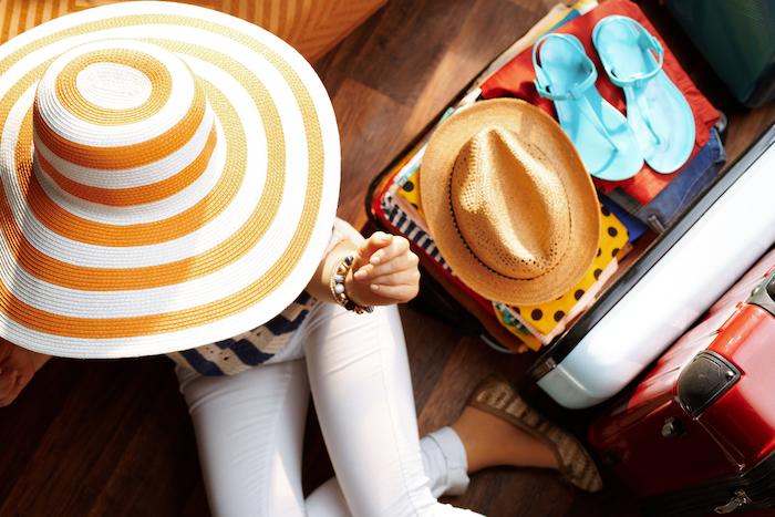 Upper view of modern woman in white pants and striped blouse with big summer hat near open travel suitcase packing for summer holiday at modern home in sunny summer day.