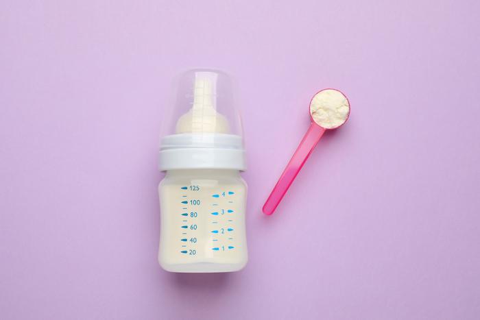 Feeding bottle with infant formula and powder on violet background, flat lay