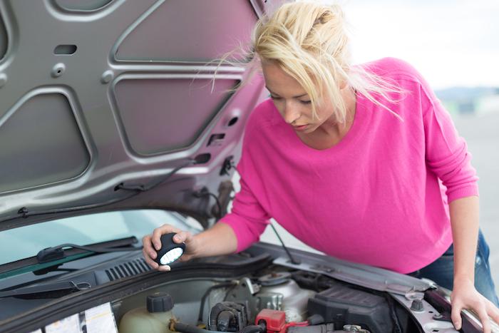 Self-sufficient confident modern young woman inspecting car engine with a flashlight in her hand.