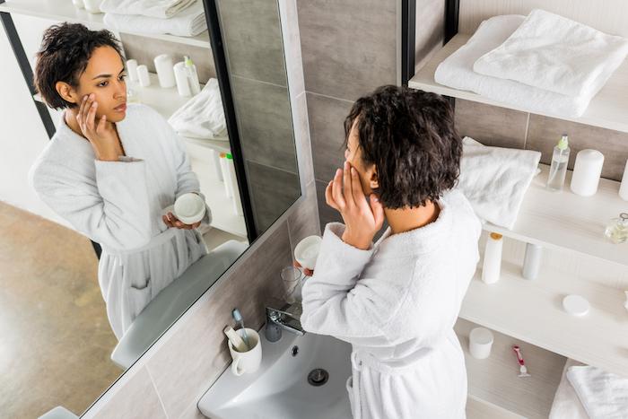 African american woman applying cosmetic anti-wrinkle cream on face and looking in mirror in bathroom