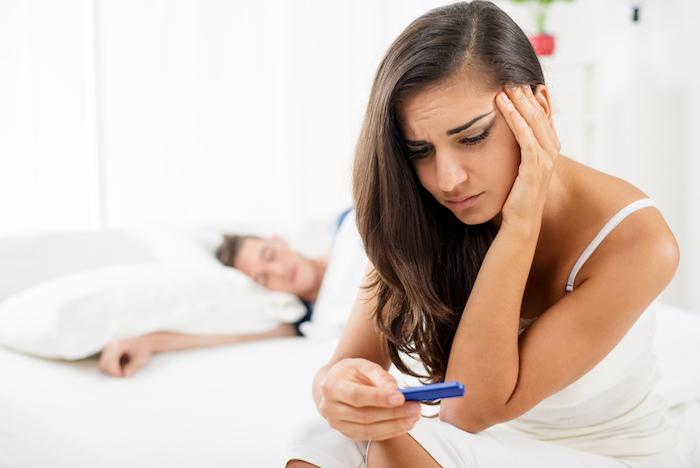 Young woman sitting on the bed with a sad expression on her face looking at pregnancy test. In the background is her partner who is sleeping in bed.