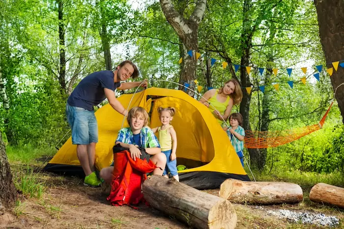 Portrait of happy young parents and three age-diverse kids putting up a tent on camping trip in the woods