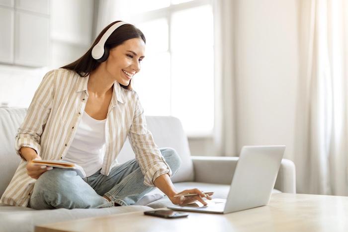 Online Education. Smiling Young Woman In Wireless Headphones Studying With Laptop At Home, Happy European Female Using Computer For Distance Learning While Sitting On Couch In Living Room