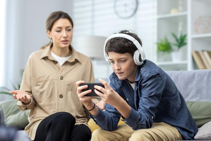 How to Protect Children from Gambling in Online Games and Loot Boxes. Mother quarrels with son, teenage boy in headphones ignores woman and plays video games on phone, family at home in living room sitting on sofa.