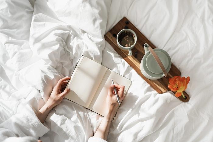 The Joy of Slow Living. High above view of person making notes using notebook and pen, kettle and morning coffee, romantic mood, cozy bedroom interior design, home decor, flat lay