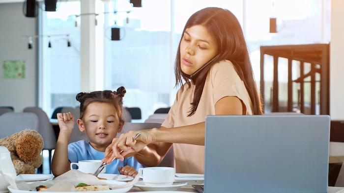 Asian businesswoman mom multitasking, gives pizza slice to toddler child sitting at table with tea and grey laptop close-up while talking on the phone at the same time
