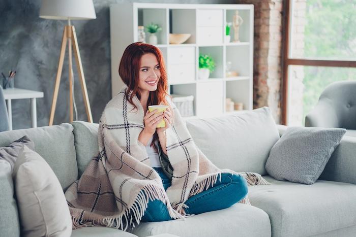 Young ginger girl sit sofa wrapped blanket drink tea cacao dressed khaki garment spacious house indoors room.