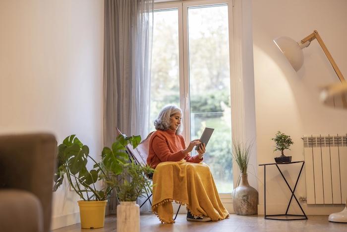 Old gray-haired woman resting at home with a tablet in her hand, by the window. take a blanket because it is cold. home life.