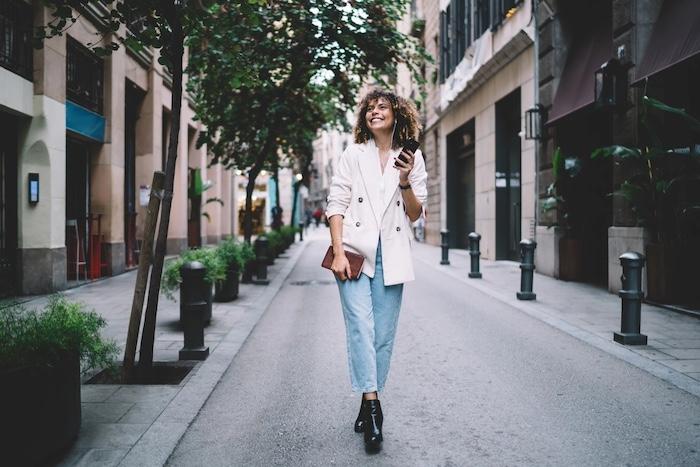 Everyday Personal Style Ideas That Balance Comfort and Fashion. Cheerful hipster girl in electronic earphones enjoying music playlist from smartphone application, joyful Caucaisan tourist received funny audio message using 4g internet on cellphone technology