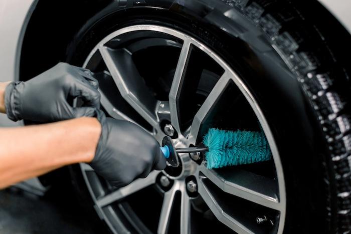 Auto wash service. Cropped close up image of male hands in black protective gloves, cleaning alloy wheels rims of luxury car with a special brush for cast wheels in a vehicle detailing workshop.